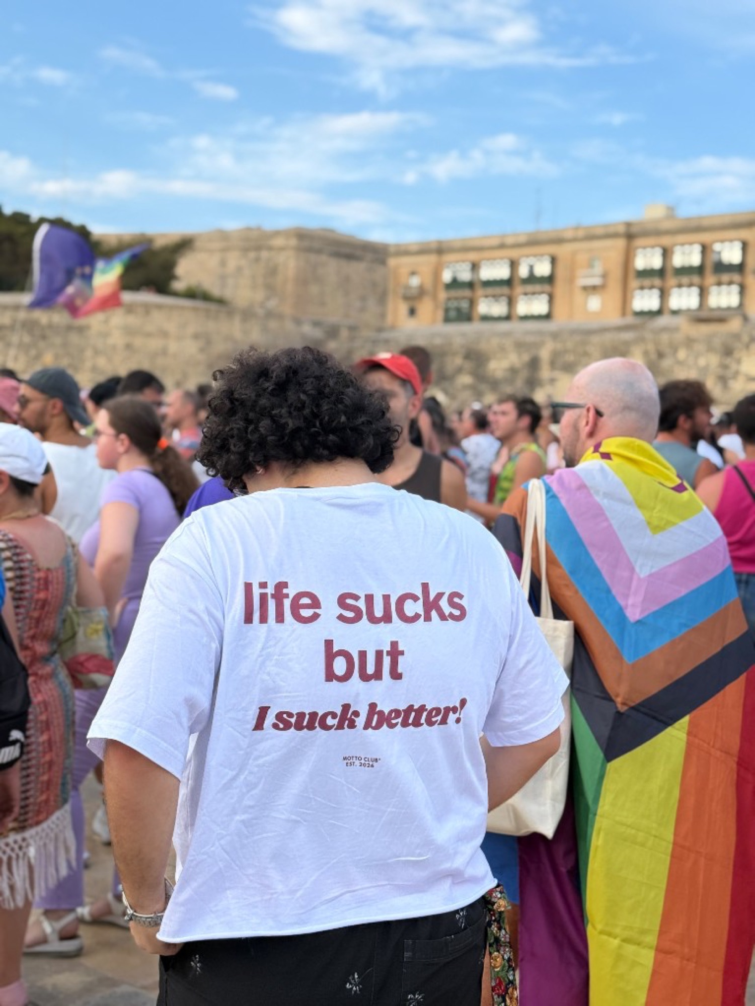 White Motto Club tee in a Valletta crowd. The back print reads life sucks but I suck better.